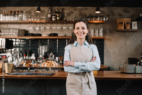 Carta da parati smiling barista in apron standing with crossed arms near bar counter
