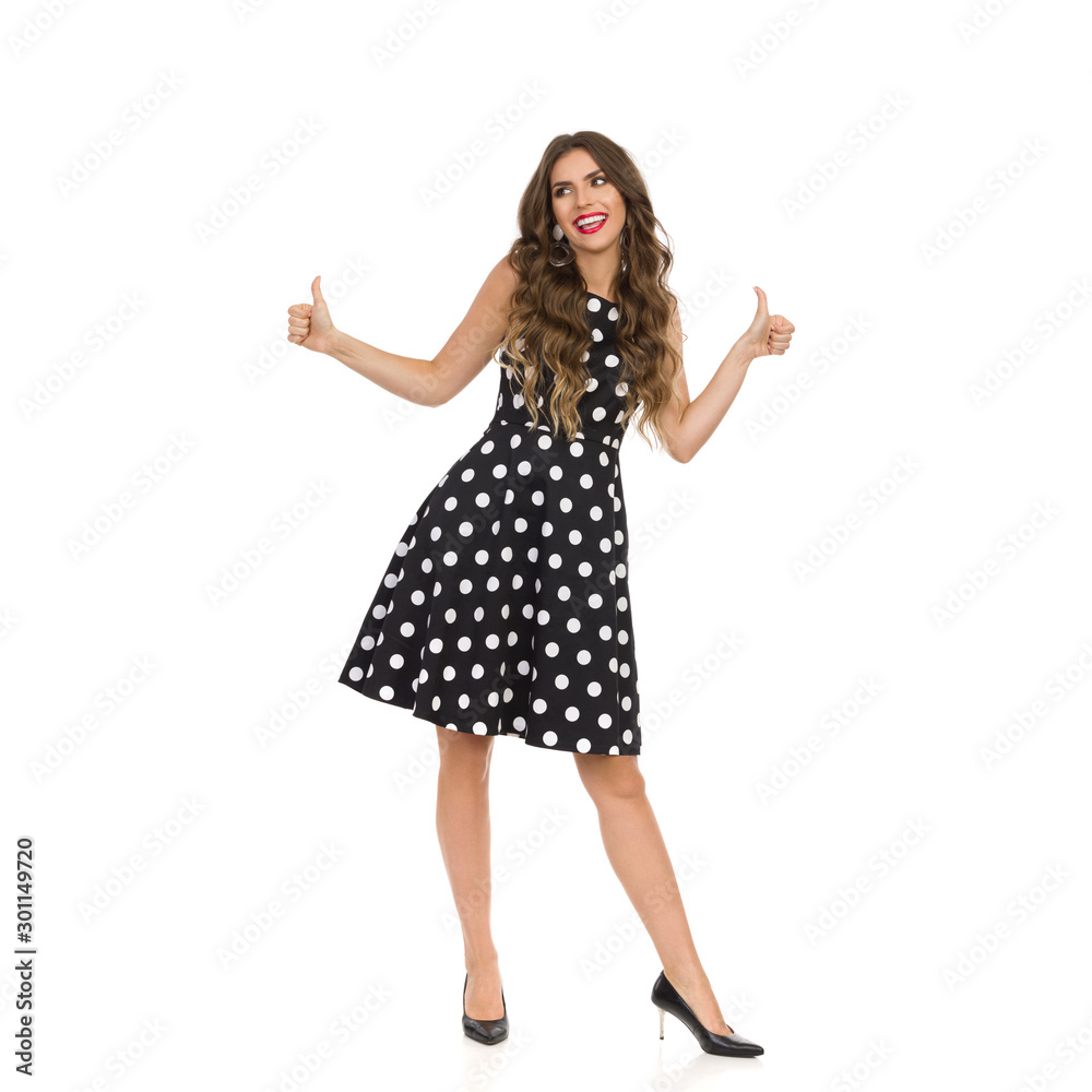 Beautiful Young Woman In Black Cocktail Dress In Polka Dots And High Heels Is Showing Thumbs Up And Talking. Beautiful Young Woman In Black Cocktail Dress In Polka Dots And High Heels Is Showing Thumbs Up And Talking.
