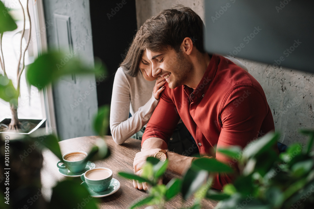 selective focus of green leaves and smiling romantic young couple hugging and drinking coffee in coffee shop - obrazy, fototapety, plakaty 