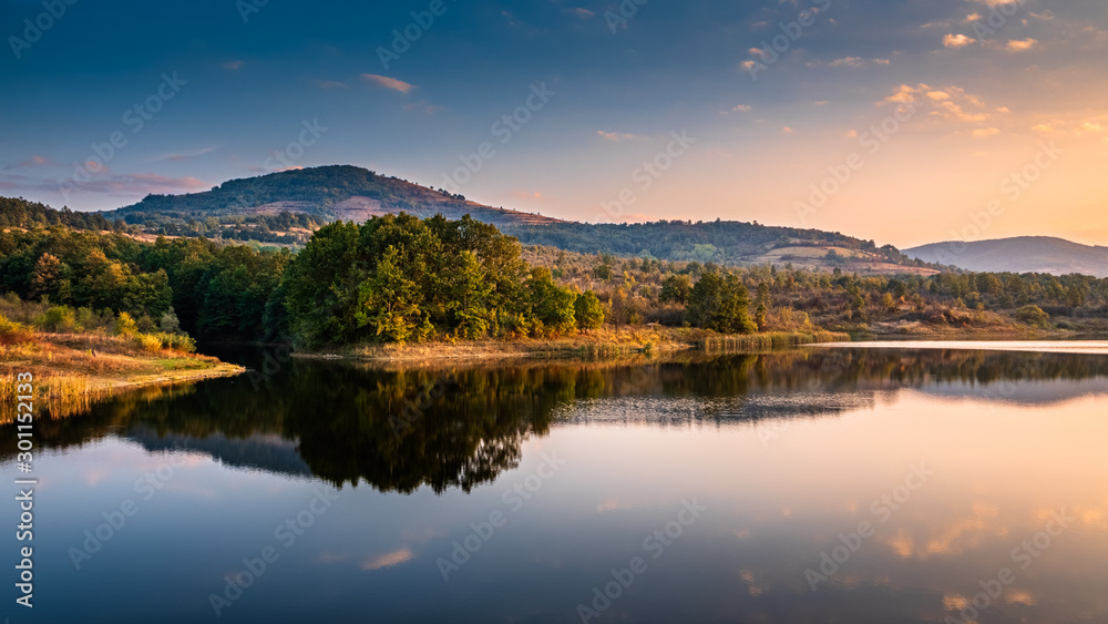 A beautiful landscape of a still water lake with natural reflections of forest hills shore in the foreground, mountain peaks and hills in the background, clear blue sky with just a few smooth clouds a