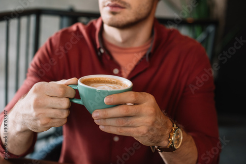 cropped view of young man with cup of tasty cappuccino in coffee shop