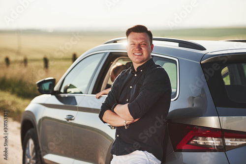 Man posing for the camera. Leaning on the car. Daughter inside automobile behind
