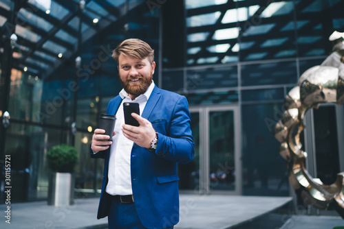 Half length portrait of cheerful male entrepreneur with takeaway cup with tasty caffeine beverage using cellphone application for checking financial news on website, concept of business people