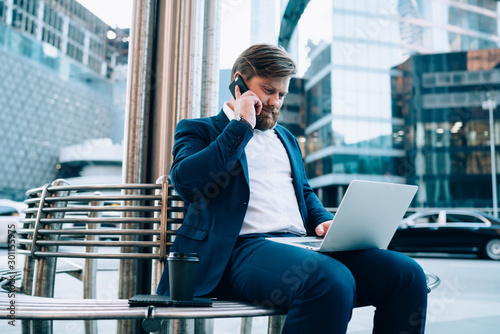 Male boss in elegant suit making notes in laptop application during mobile conversation with colleague, serious businessman talking with service operator via smartphone gadget for checking balance