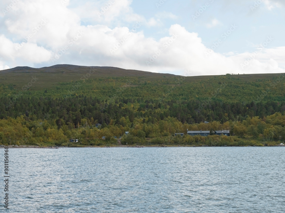 view on sami hut in Saltoluokta from river Lulealven with green mountains, birch tree and blue sky white clouds