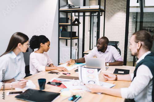 Multicultural team of creative male and female designers teamworking on developing common project during brainstorming, experienced business people in formal wear discussing financial reports