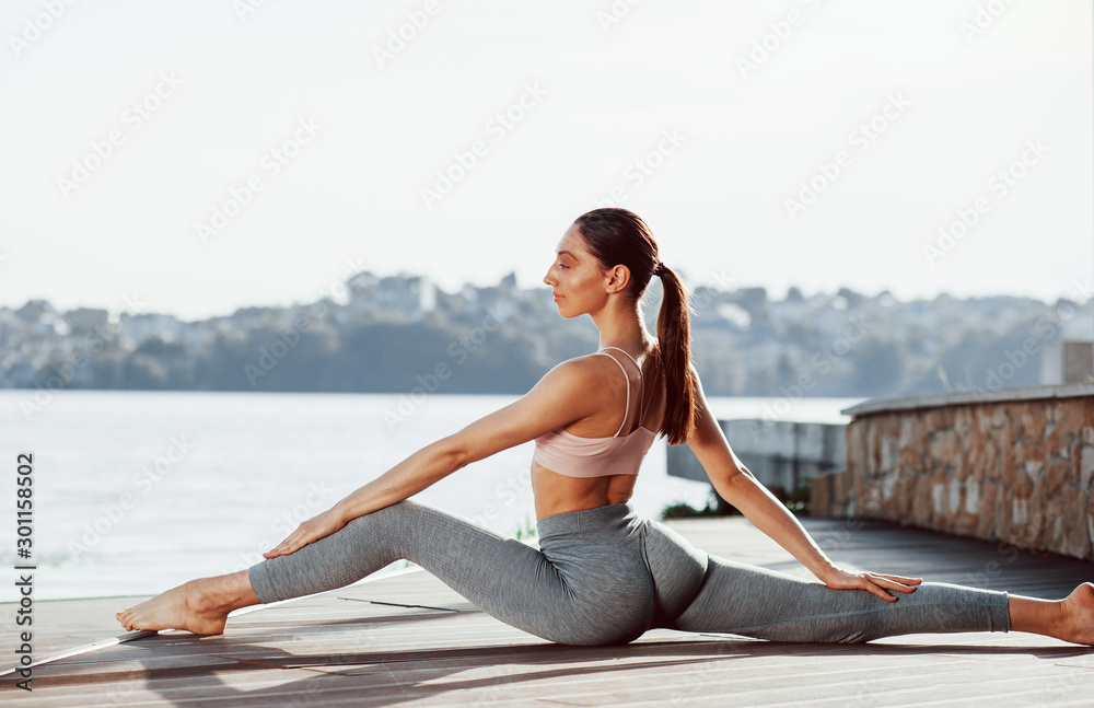 Feeling calm. Young woman with slim type of body does exercises against lake