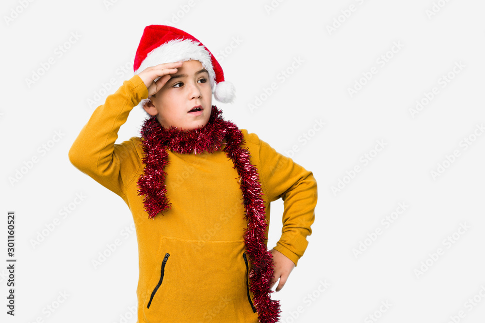 Little boy celebrating christmas day wearing a santa hat isolated looking far away keeping hand on forehead. Little boy celebrating christmas day wearing a santa hat isolated looking far away keeping hand on forehead.