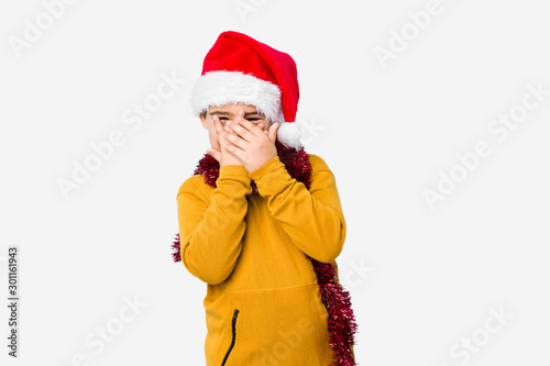 Little boy celebrating christmas day wearing a santa hat isolated blink through fingers frightened and nervous.