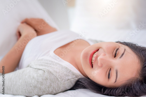 Beautiful young pretty Asian woman wake up and make happy smile with white shirt at the white bed in the morning.
