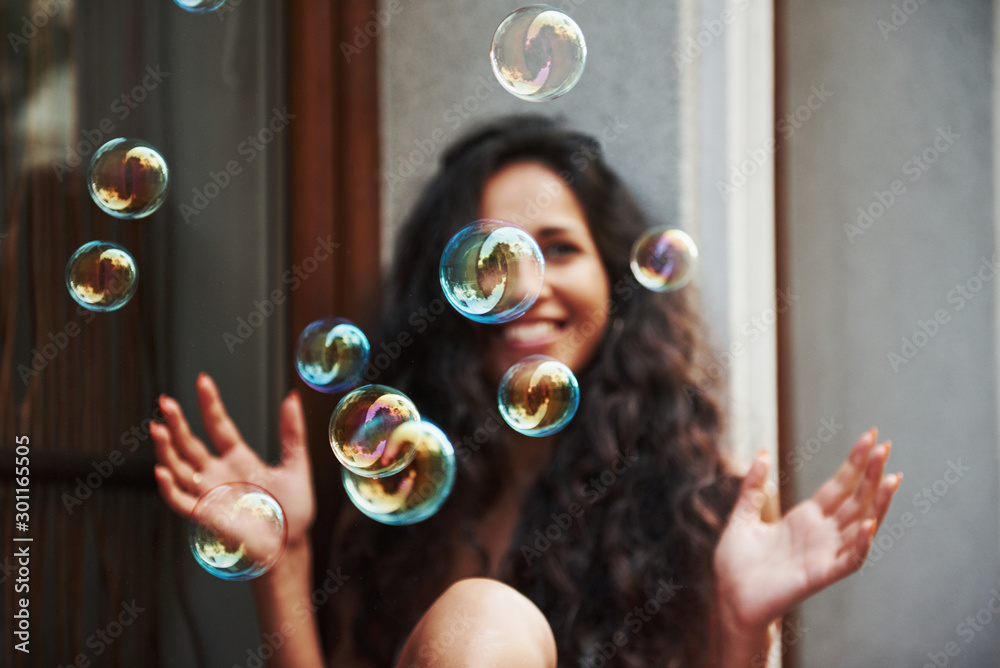 Cute girl plays with bubbles like a child. Beautiful woman with curly black hair have good time in the city at daytime - obrazy, fototapety, plakaty 