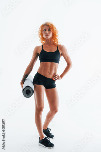 Holds grey colored roll mat in hand. Redhead female bodybuilder is in the studio on white background