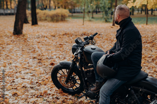 Back view of bearded male biker looks thoughtfully somewhere into distance, poses on black motorbike, holds protective helmet, spends leisure time in autumnal park, copy space for your advertising