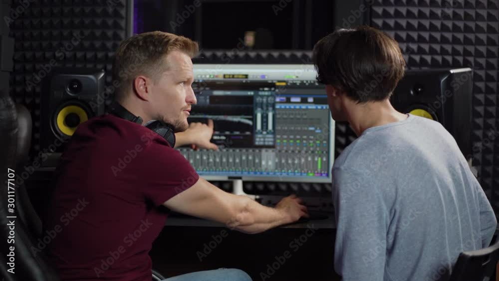Zoom in back view medium shot of sound engineer and music producer using computer with professional software and discussing recorded song in sound recording studio Zoom in back view medium shot of sound engineer and music producer using computer with professional software and discussing recorded song in sound recording studio