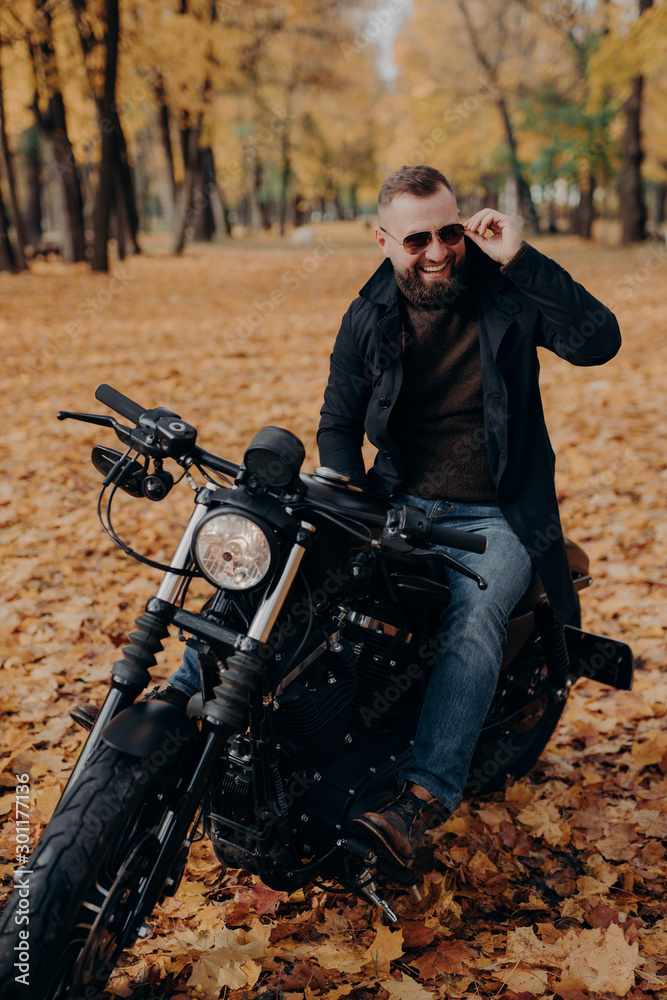 Image of cheerful male motorcyclist smiles joyfully, keeps hand on shades, sits on fast bike, has fun outdoor, enjoys traveling by motorbike, spends free time in autumn park. Travel concept.