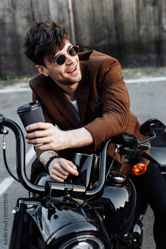 young man sitting on motorcycle, smiling, looking away and holding paper cup of coffee