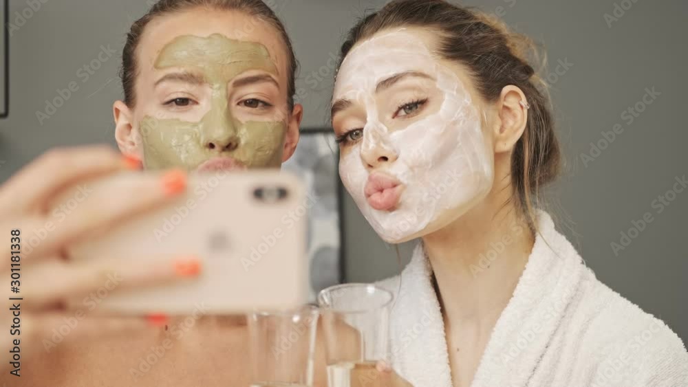 Two pretty smiling girls friends with facial mask on their faces making selfie by smartphone at home