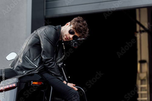 young motorcyclist in sunglasses and leather jacket while sitting on motorcycle near garage