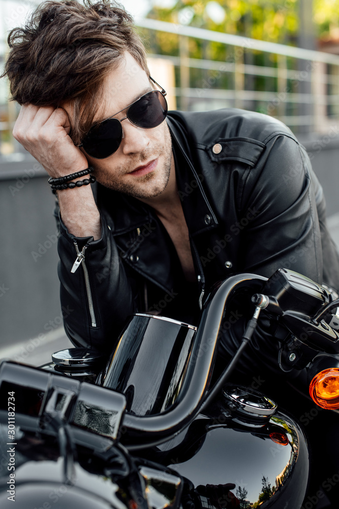 handsome young motorcyclist in sunglasses holding head with hand and looking away while sitting on motorcycle
