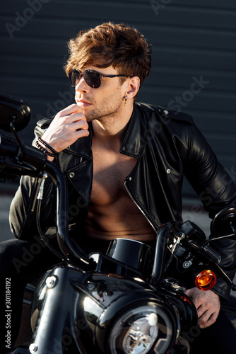 young man in sunglasses and black leather jacket with naked torso looking away and sitting on motorcycle