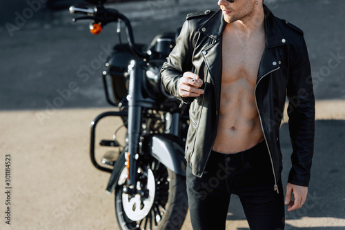 cropped view of man with muscular torso in leather jacket looking away and smoking cigarette