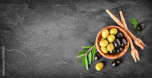 Fresh olives in wooden bowl on dark stone table. Black and green olive with pickers or sticks from top view. Banner or panorama concept.