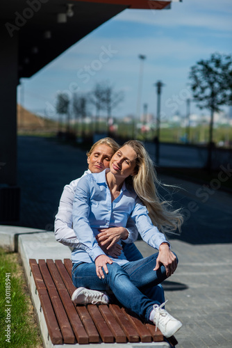 Two surprisingly similar women are sitting on a bench in an embrace and smiling. A middle-aged woman and her elderly mother. Beautiful well-groomed mother and daughter family look.