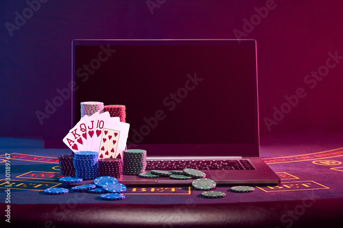 Chips piles and playing cards on laptop standing on blue cover of a table. Black background. Gambling entertainment, poker, casino concept. Close-up.