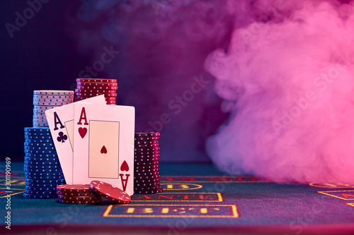Two aces, hearts and clubs, leaning on colored chips piles on blue cover of playing table. Black, smoke background, red and blue backlights. Casino.