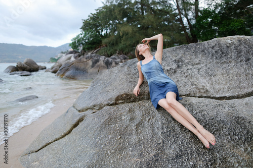 Young caucasian woman lying on stones on sea shore in morning, wearing jeans sundress. photo