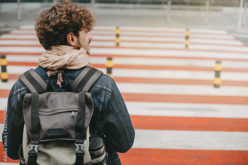 Obraz premium Young attractive man with curly hair stand in front of crosswalk and look to right. Carrying backpack. Walking to airport or railway station. Going abroad or on vacation.