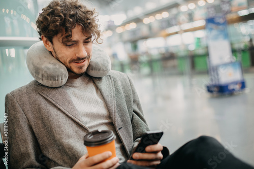 Picture of young man in suit sitting in waiting room and use phone in hands. Wait for train or airplane. Hold cup of coffee in hands. Pillow around neck. Calm and concentrated. Manager travel abroad.