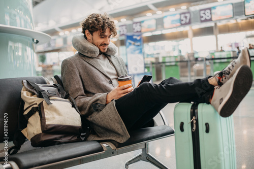 Young businessman sit alone and wait for flight. Hold phone in hands and cup of coffee. Flight pillow around neck. Expert or executive manager travel abroad. Waiting room.
