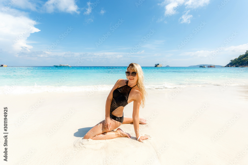 Blonde young woman in black swimsuit sitting on side at beach. Beautiful tanned girl lying on sand. Attractive girl wearing glamour swimwear on tropical beach while sunbathing