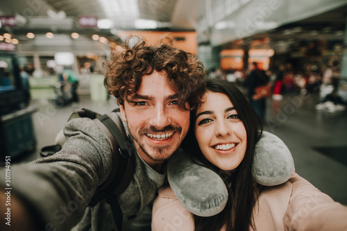 Beautiful young couple posing on camera together. Look stragight and smile. Inside airport. Guy hold camera with hand. Travel pillow around her neck. Trip or vacation.