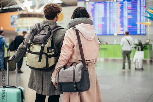 Back view of couple in airtport. Carry purses and suitcase. Ready for trip or vacation. Carry backpacks as well. Look at travel board with flights information. Stand together.