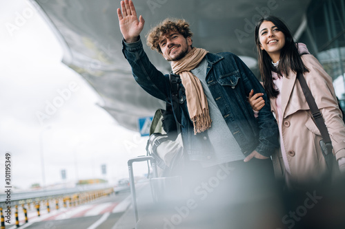 Close up and cut view of young couple after vacation trying to catch taxi cab. Guy wave with hand. Georgian woman hold his amother hand and look straight with smile. Daylight. Stand outside airport. photo