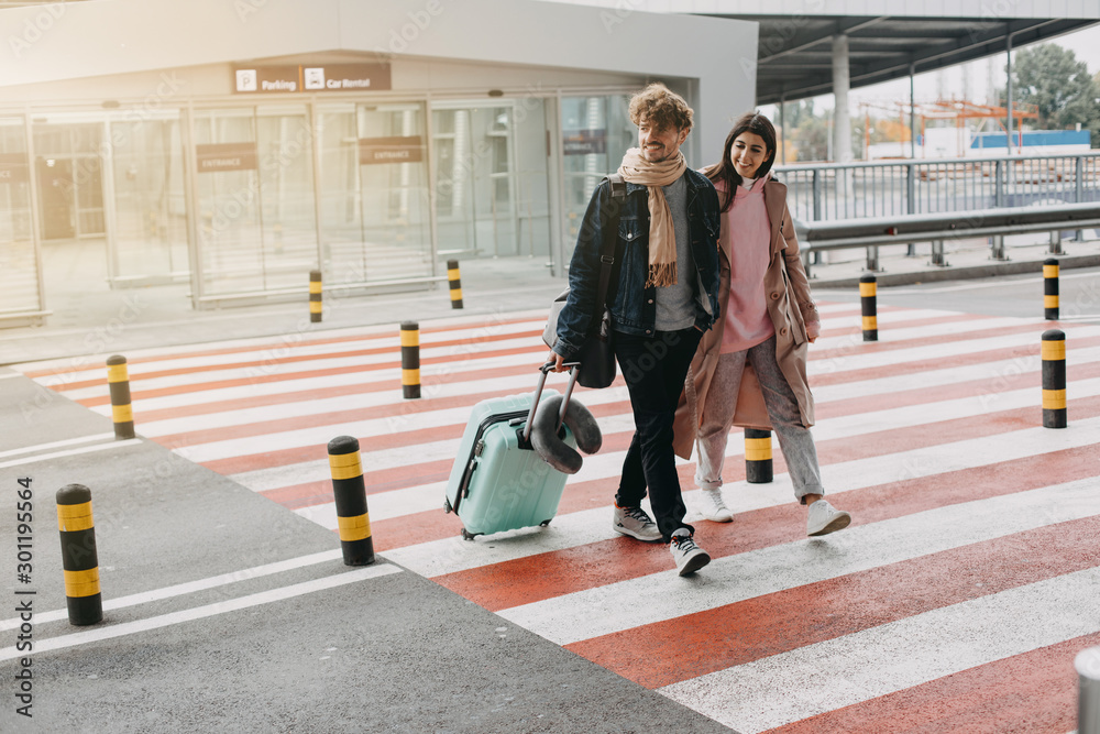 Picture of young man and woman walk on crosswalk together. Guy carry suitcase. Goint to travel abroad. Vacation or trip. Sunlight on picture. In rush to airport.