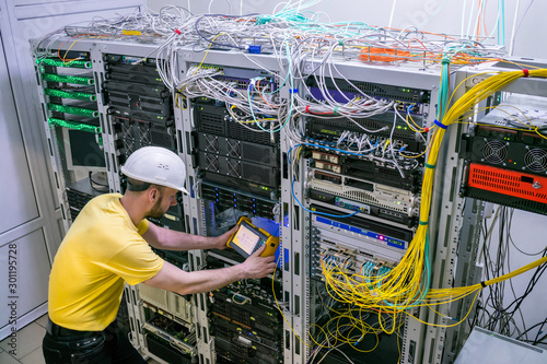 The engineer in a white helmet measures the level of the optical signal in the server room of the data center. A technician diagnoses a problem area in a telecommunication network.