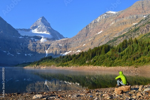 Young man is sitting on the banch of lake in frosty morning. Mount Assiniboine is towering above him. Mount Assiniboine Provincial Park, Canada.