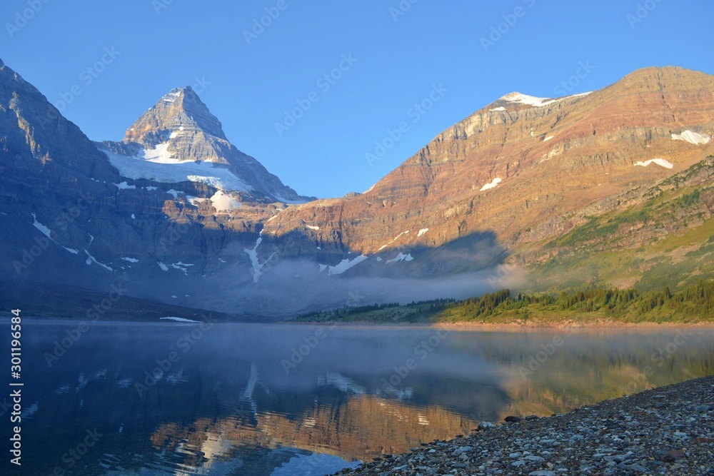 Cold sunny morning in Assiniboine Provincial Park. Lake in front with rolling fog, Mount Assiniboine covered with snow in towering over whole scenery.
