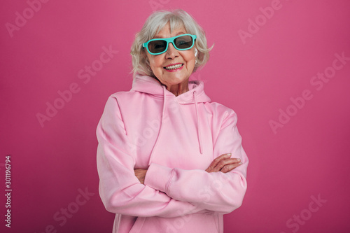 Cheerful, positive and cool old woman wear pink hoody and green with black sunglasses. Hands crossed and psoing on camera. Stand alone. Isolated over pink bakground.