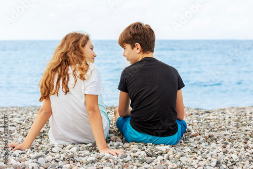 Couple of beautiful teens, first love. Guy hugs a girl sitting on the pebble beach next to each other and looking at the sea. Shoot from behind