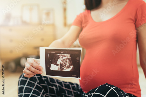 Pregnant brunette sitting on floor alone in room. Hold ultrasonic picture with one hand. Waiting for childbirth. Future baby in belly. Cut view. photo