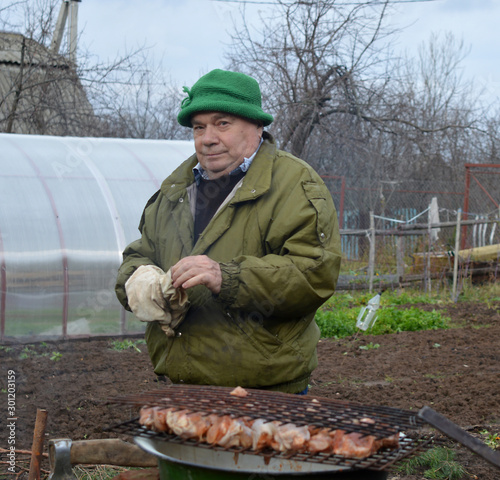 An elderly man cooks food over a fire in the garden in autumn and laughs © Елена Пахмутова