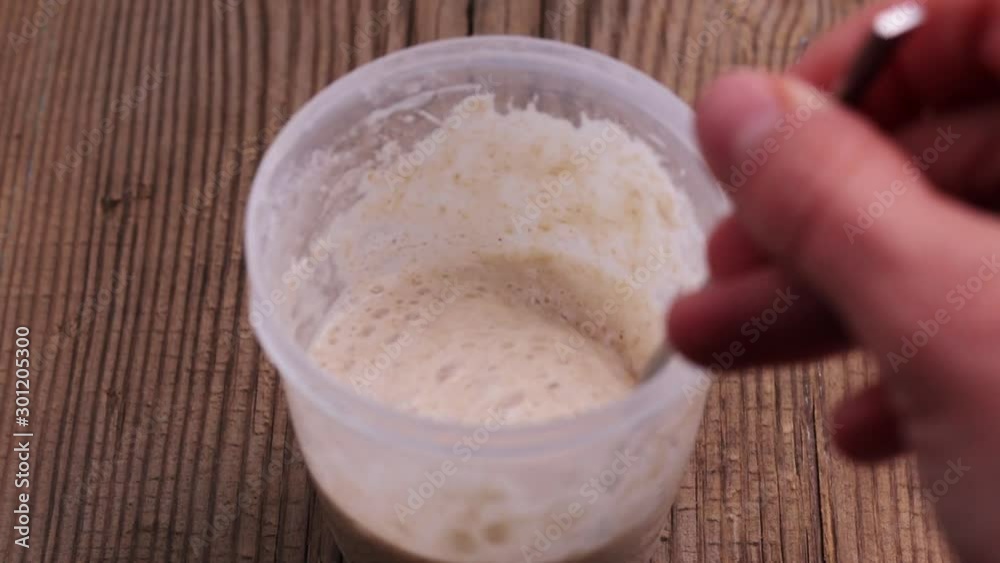 Homemade sourdough starter with lots of bubbles in small jar is mixing with tea spoon on old textured wooden table. Bakery, baking bread and fermentation Homemade sourdough starter with lots of bubbles in small jar is mixing with tea spoon on old textured wooden table. Bakery, baking bread and fermentation