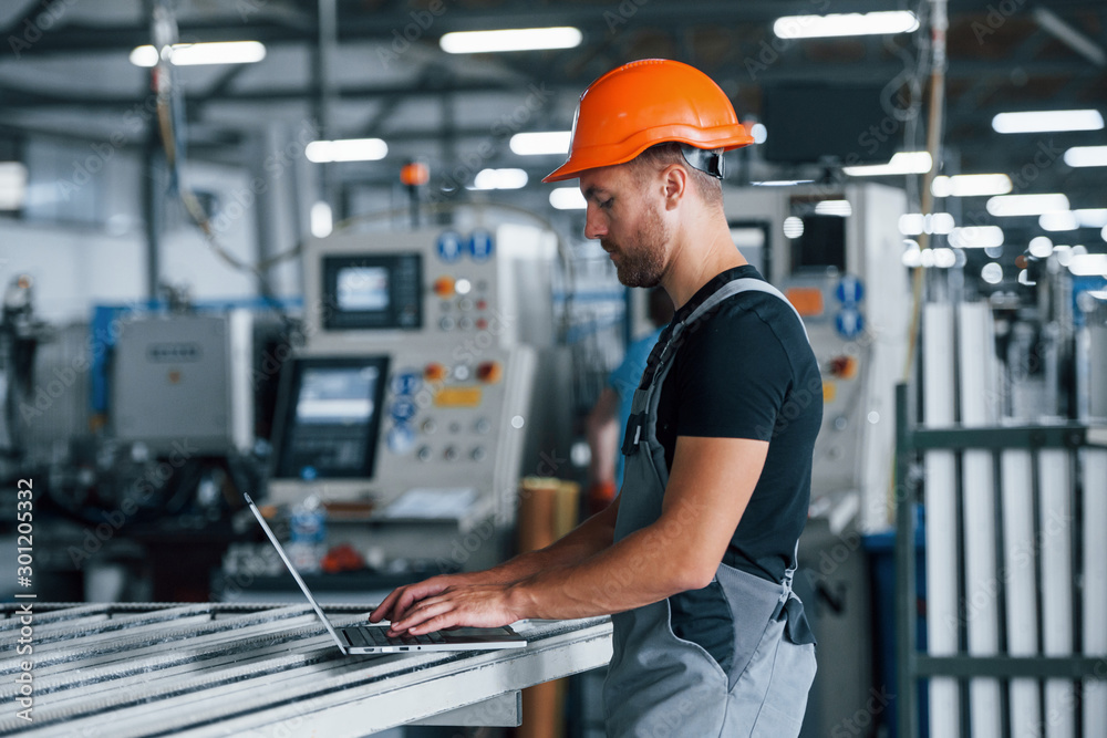 Modern laptop. Typing on keyboard. Industrial worker indoors in factory. Young technician with orange hard hat Modern laptop. Typing on keyboard. Industrial worker indoors in factory. Young technician with orange hard hat