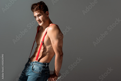 Фотография shirtless man in suspenders looking at camera and standing near white chair isol