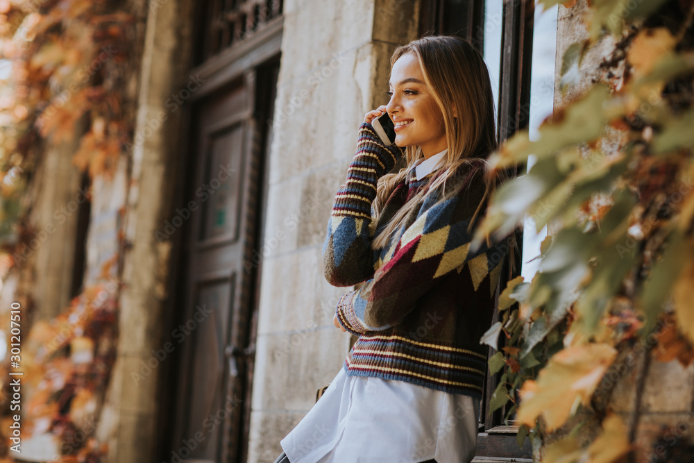 Fototapeta premium Pretty young woman standing with mobile phone on street at autumn day