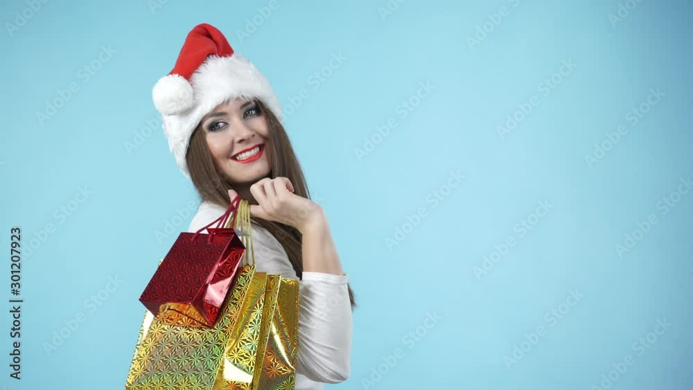 Happy adorable young woman in santa claus helper hat holding many shopping bags, gifts packaging, on blue. Christmas holidays Happy adorable young woman in santa claus helper hat holding many shopping bags, gifts packaging, on blue. Christmas holidays